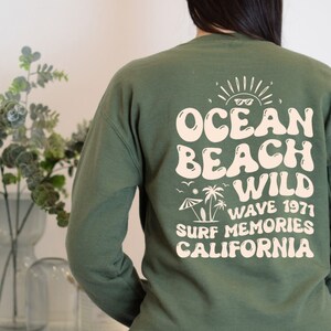 May include: A military green sweatshirt with a retro-style graphic print on the back. The print features the text "Ocean Beach Wild Wave 1971 Surf Memories California" in a cream color.