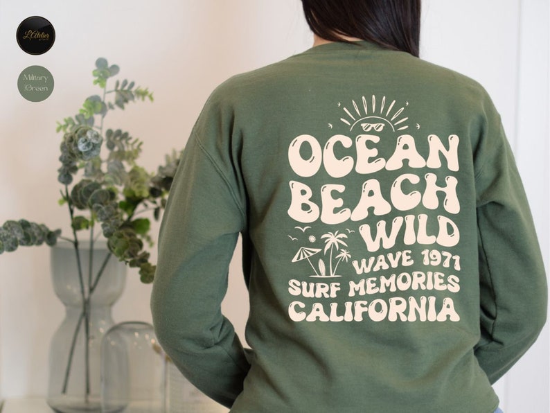 May include: A military green sweatshirt with a retro-style graphic print on the back. The print features the text "Ocean Beach Wild Wave 1971 Surf Memories California" in a cream color.