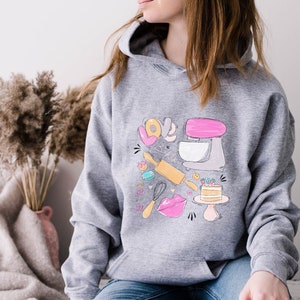 May include: A gray hoodie with a graphic design of a pink stand mixer, rolling pin, whisk, cake, and the words "Love & Bake".