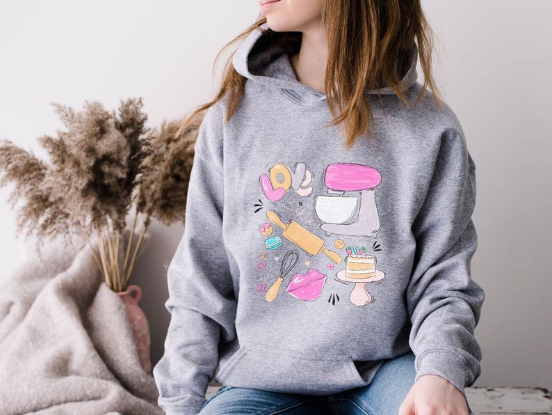 May include: A gray hoodie with a graphic design of a pink stand mixer, rolling pin, whisk, cake, and the words "Love & Bake".