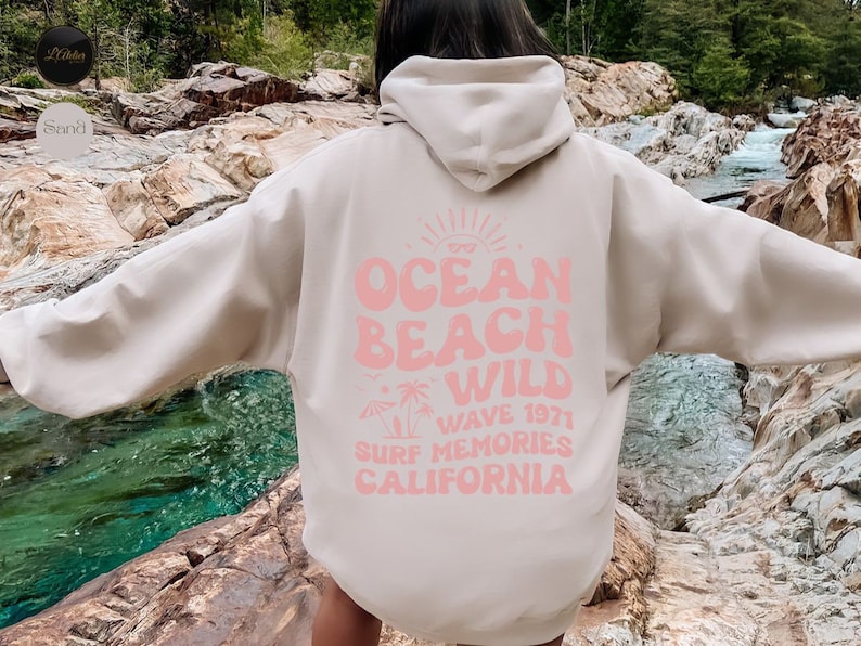 May include: A light beige hoodie with a pink graphic print that says "Ocean Beach Wild Wave 1971 Surf Memories California".
