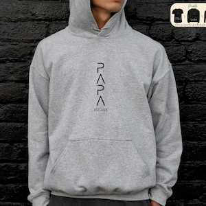 May include: A gray hooded sweatshirt with a black graphic design that reads "PAPA" with a triangle symbol between each letter and "EST 2025" below the graphic.