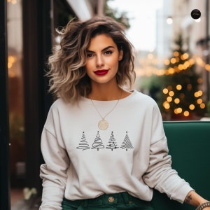 May include: A white sweatshirt with a black outline of four Christmas trees. The trees are stylized and have a simple design. The sweatshirt has a gold necklace with a round pendant.