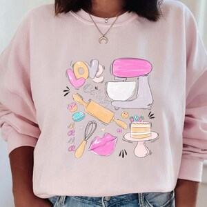 May include: Pink sweatshirt with a graphic design of a kitchen mixer, rolling pin, whisk, cake, and the words "Love Baking".