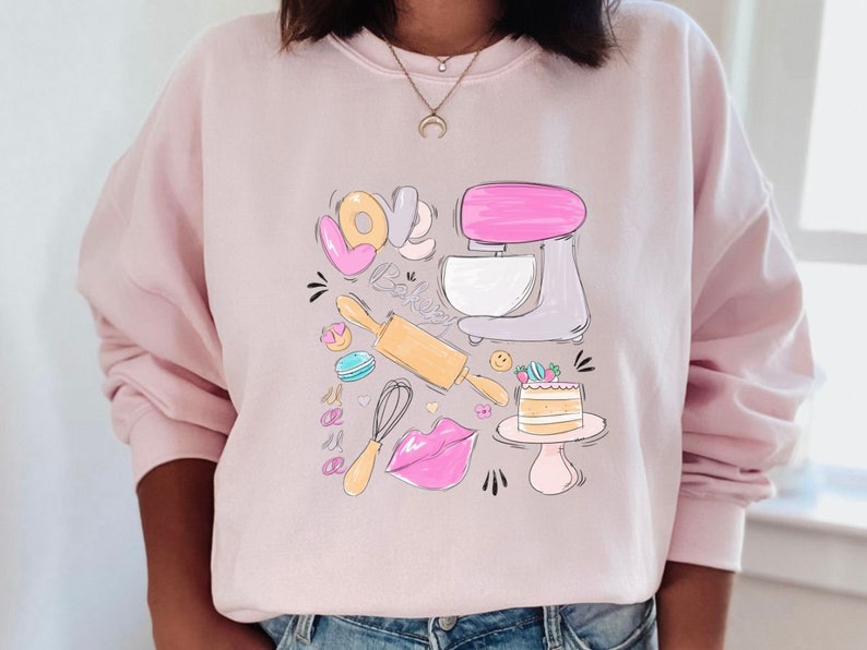 May include: Pink sweatshirt with a graphic design of a kitchen mixer, rolling pin, whisk, cake, and the words "Love Baking".