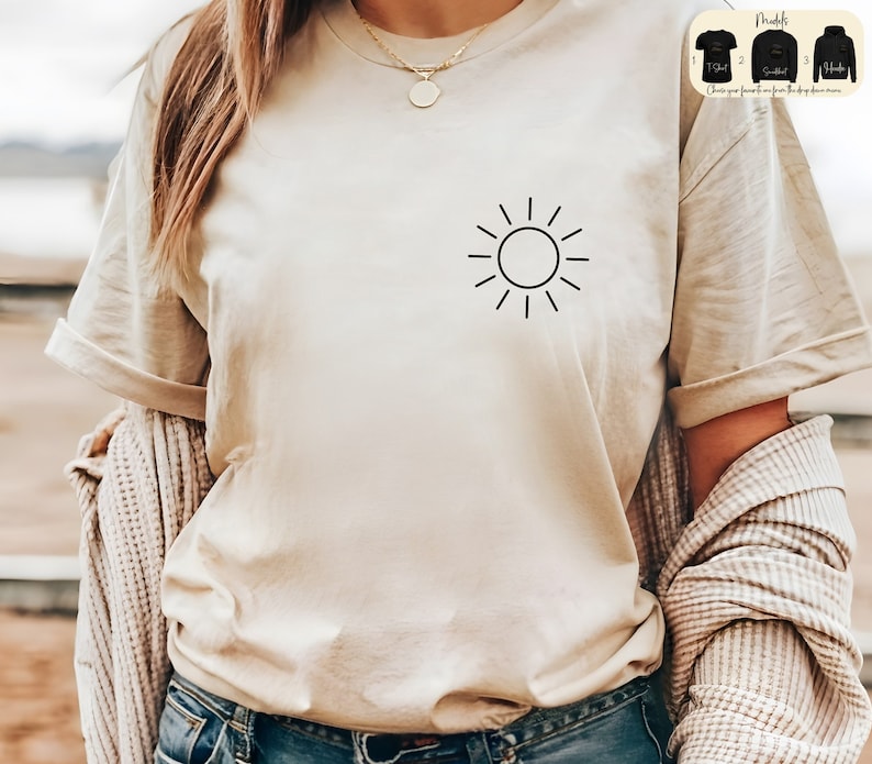May include: A beige t-shirt with a simple black sun graphic on the left chest. The shirt is being worn by a person with long brown hair.