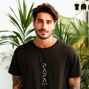 May include: A man wearing a black t-shirt with a white graphic design that says "PAPA EST 2025".
