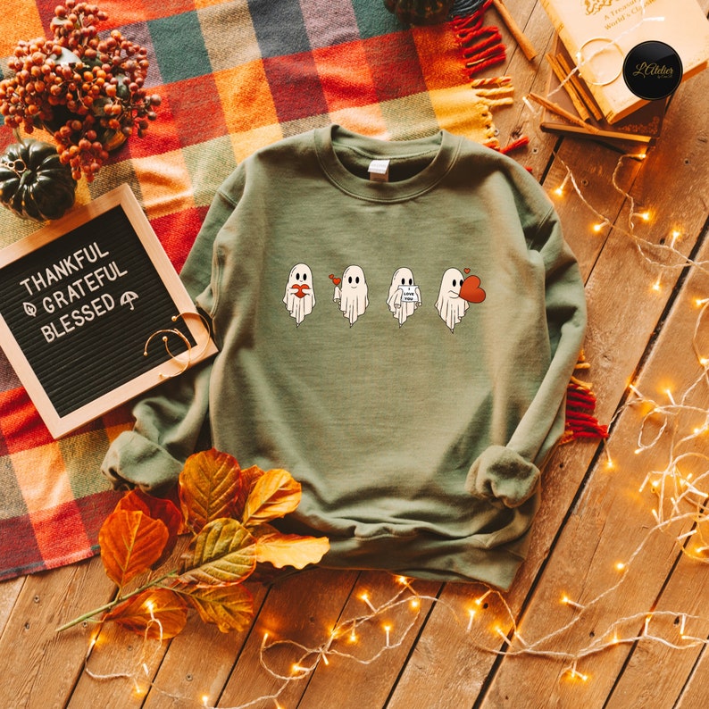 May include: A green sweatshirt with a graphic of four ghosts. The ghosts are wearing different expressions, including a heart, a kiss, a note that says "Love You", and a red heart. The sweatshirt is on a wooden surface with fall leaves and string lights.