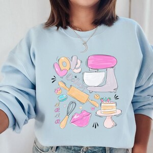 May include: A light blue sweatshirt with a graphic design of a kitchen mixer, rolling pin, whisk, and a cake. The graphic also includes the words "Love Bake".