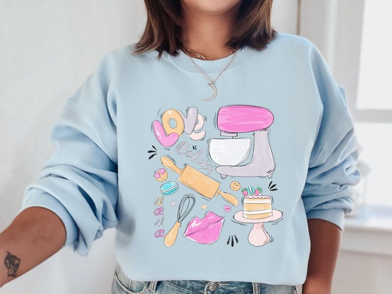 May include: A light blue sweatshirt with a graphic design of a kitchen mixer, rolling pin, whisk, and a cake. The graphic also includes the words "Love Bake".