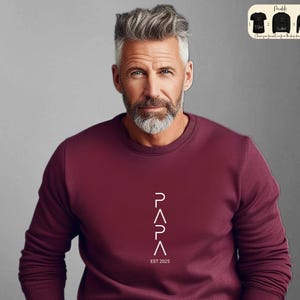 May include: A maroon sweatshirt with a white graphic design that reads "PAPA" with a triangle shape for each letter. The text "EST 2025" is below the graphic.