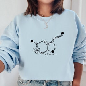 May include: A light blue sweatshirt featuring a black floral design of the chemical structure of serotonin. The text "Serotonin" is written below the design.