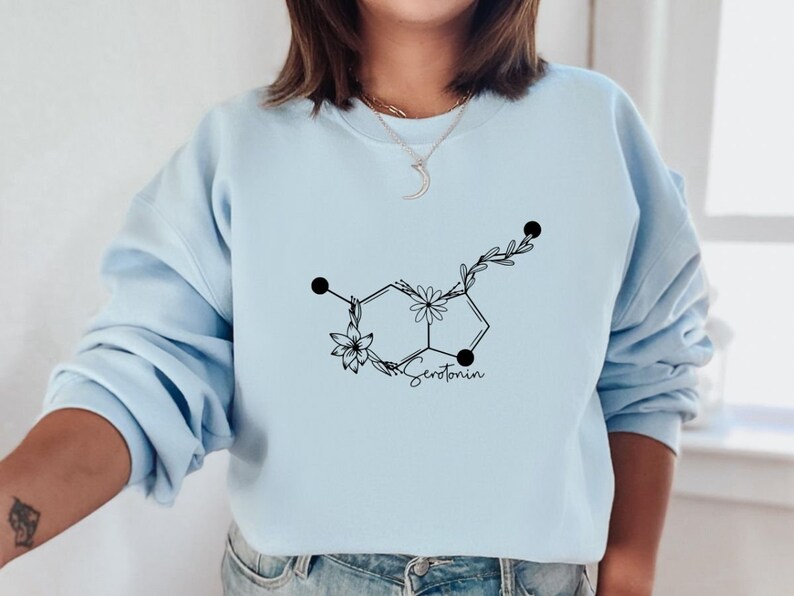 May include: A light blue sweatshirt featuring a black floral design of the chemical structure of serotonin. The text "Serotonin" is written below the design.