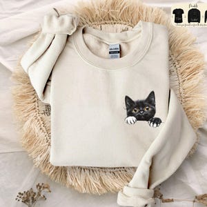 Puede incluir: Sudadera de color crema con un gráfico de un gato negro con ojos amarillos y patas blancas asomando. La sudadera tiene cuello redondo y mangas largas. La imagen también muestra tres modelos diferentes de la camisa.