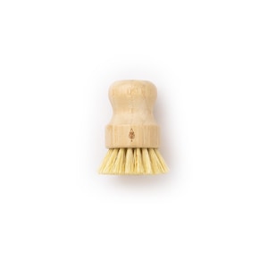 May include: A small, round, wooden-handled brush with natural bristles. The brush has a small, brown logo on the handle.