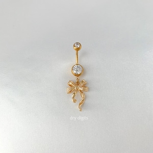 May include: A gold-colored belly button ring with a bow charm. The ring features a clear, round gemstone at the top and a larger gemstone in the center. The bow charm hangs below the center gemstone. The text "dry digits" is at the bottom.