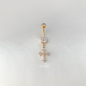 May include: Gold-colored belly button ring with a dangling cross pendant. The ring features a large, clear gemstone at the center and a smaller gemstone at the top. The cross pendant is adorned with multiple small, clear gemstones. The text "dry digits" is visible.