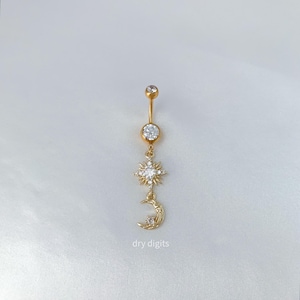 May include: Gold-coloured belly button ring with a celestial design. The jewellery features a large, clear gem at the top, a sunburst design, and a crescent moon charm. The text "dry digits" is at the bottom.