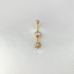 Könnte beinhalten: Goldfarbener Bauchnabelring mit einem Sonnenstrahl-Anhänger. Der Ring hat einen klaren Edelstein oben und einen größeren Edelstein in der Mitte. Der Sonnenstrahl-Anhänger hat eine kleine Perle in der Mitte. Der Text "dry digits" steht unten.