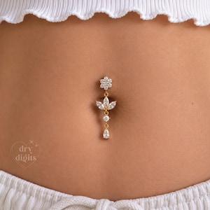 May include: A gold-toned navel ring featuring a flower-shaped top set with clear stones, a central leaf-like arrangement of marquise-cut stones, a single round stone, and a pear-shaped drop stone. The jewelry is positioned in a navel piercing.