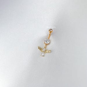 Pode incluir: Piercing de umbigo dourado com um pingente de anjo. O anjo tem pequenas asas e um corpo de pedra preciosa transparente. O piercing apresenta uma gema grande e transparente e uma gema menor no topo.