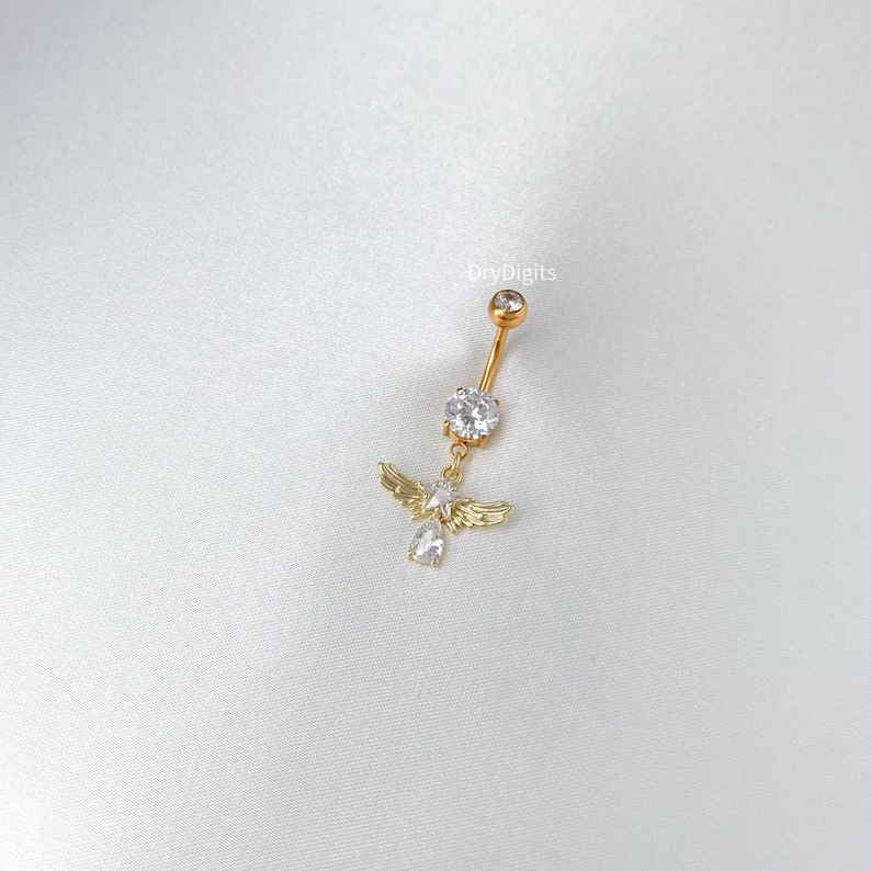 Pode incluir: Piercing de umbigo dourado com um pingente de anjo. O anjo tem pequenas asas e um corpo de pedra preciosa transparente. O piercing apresenta uma gema grande e transparente e uma gema menor no topo.