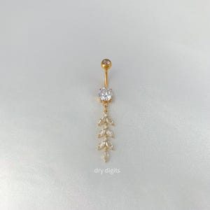 May include: Gold-colored belly button ring with a dangling leaf design. The top features a small, clear gem, with a larger gem in the center. The dangling portion has three leaf-shaped gems. The text "dry digits" is at the bottom.