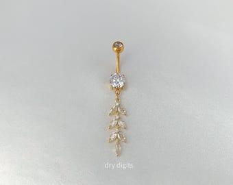 May include: Gold-colored belly button ring with a dangling leaf design. The top features a small, clear gem, with a larger gem in the center. The dangling portion has three leaf-shaped gems. The text "dry digits" is at the bottom.