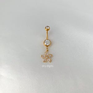 May include: A gold-colored belly button ring with a flower charm. The ring features a clear gem at the top and a larger gem in the center. The flower charm is made of clear gems. The text "dry digits" is visible.