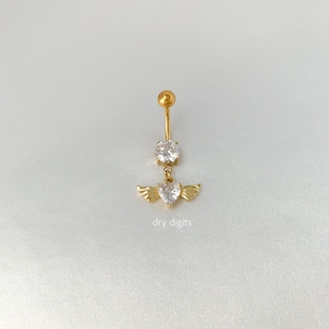 May include: A gold-colored belly button ring with a clear, round gem at the top and a heart-shaped gem with small wings at the bottom. The text "dry digits" is printed at the bottom of the image.