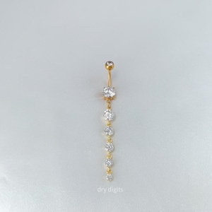 May include: Gold-toned belly button ring with a dangling design. The jewelry features a small top ball and a series of six clear, round gemstones of varying sizes. The text "dry digits" is at the bottom.