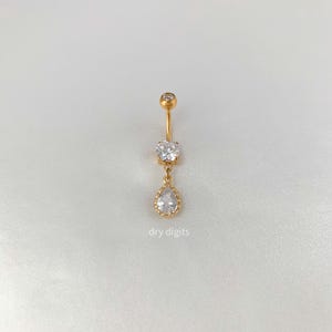 以下が含まれることがあります： 3つの透明で輝く宝石が付いた、金色のへそピアス。上と真ん中の石は丸く、下の石は涙の形をしています。「dry digits」の文字が下部に印刷されています。