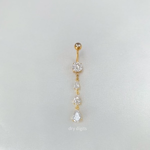 Puede incluir: Un piercing para el ombligo de color dorado con un diseño colgante. El anillo presenta una pequeña gema redonda en la parte superior, seguida de una gema redonda más grande y tres gemas más pequeñas en forma de lágrima. El texto "dry digits" está en la parte inferior.