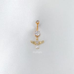 May include: Gold-colored belly button ring with a dangling angel wing design. The ring features a large, clear, round gem at the top and a smaller gem at the bottom. The angel wings and gems are encrusted with smaller, clear stones.