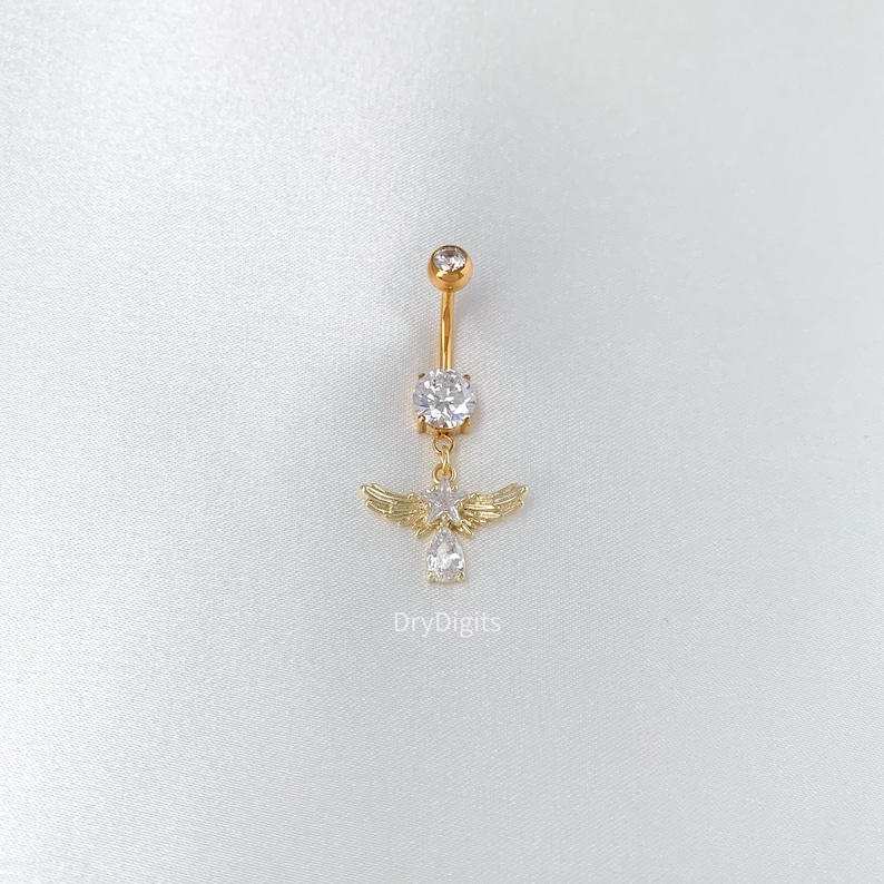 Pode incluir: Anel de umbigo dourado com um design de asas de anjo penduradas. O anel apresenta uma gema grande, transparente e redonda no topo e uma gema menor na parte inferior. As asas de anjo e as gemas s&atilde;o incrustadas com pedras menores e transparentes.