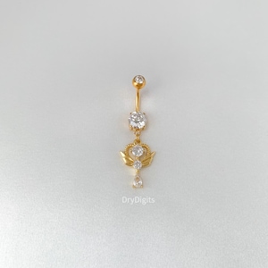 May include: Gold-coloured belly button ring with a dangling design. The jewellery features a large, clear, round gem at the top and a smaller gem at the bottom. A crown and wing design is in the middle. The text "DryDigits" is at the bottom.
