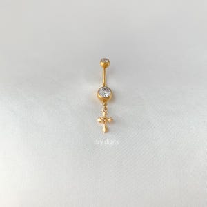 May include: Gold-coloured belly button ring with a dangling cross charm. The ring features a large, clear gemstone at the centre and a smaller one at the top. The cross charm is also adorned with small, clear gemstones. The text "dry digits" is at the bottom.