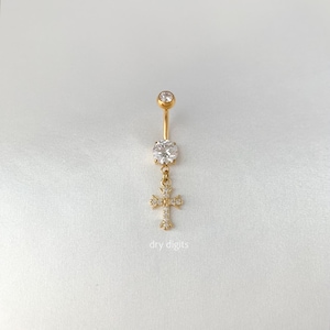 Puede incluir: Anillo para el ombligo de color dorado con un colgante de cruz. El anillo presenta una gema grande y transparente en la parte inferior y otra más pequeña en la parte superior. La cruz está adornada con múltiples gemas pequeñas y transparentes. El texto "dry digits" está en la parte inferior.