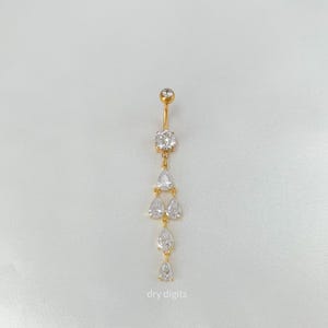 Puede incluir: Anillo para el ombligo dorado con un diseño colgante. La joya presenta una gema clara grande y redonda en la parte superior y una serie de gemas claras en forma de pera y redondas colgando debajo. El texto "dry digits" está en la parte inferior.