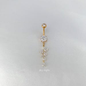 May include: A gold-coloured belly button ring featuring a cascade of clear, sparkling gemstones. The top and central stones are round, with pear and teardrop-shaped stones dangling below. The text "dry digits" is visible at the bottom.