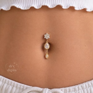 May include: A gold-toned navel ring featuring a flower-shaped top set with clear stones, a central pear-shaped drop stone, and a small marquise-shaped bottom stone. The jewellery is positioned in a navel. The text dry digits is visible in the corner.