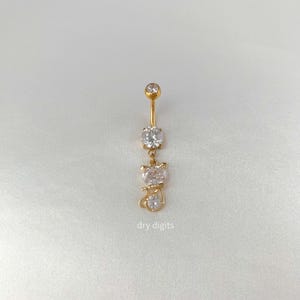 May include: A gold-colored belly button ring with three clear, sparkling gemstones. The design includes a round top stone, a larger round stone, and a cat-shaped charm with a smaller stone. The text "dry digits" is printed at the bottom.