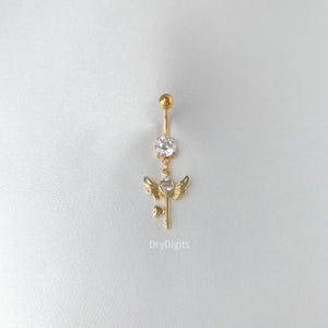 May include: Gold-colored belly button ring with a dangling charm. The charm features a key design with heart-shaped accents and small wings. A large, clear gemstone is set above the charm. The text "DryDigits" is printed on the bottom.