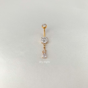 Könnte beinhalten: Ein goldfarbener Bauchnabelring mit einem hängenden Design. Der Ring hat einen großen, runden, klaren Edelstein und einen kleineren, tropfenförmigen Edelstein. Der Text "dry digits" ist unten sichtbar.