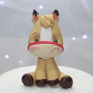 Peut inclure: Une figurine de cheval en pâte à sucre marron et blanche avec une bride rouge. Le cheval est assis les jambes croisées et a une expression mignonne et caricaturale.