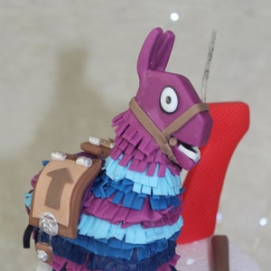 Può includere: Un topper per torta a forma di lama piñata, di colore viola e blu, con una sella marrone e una piccozza argentata. Il lama ha un occhio bianco e un cinturino marrone intorno al collo.