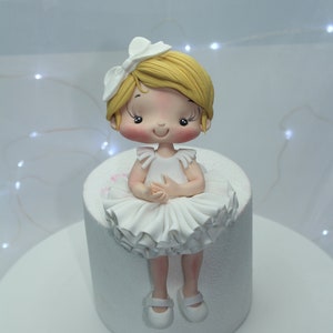 May include: A white fondant ballerina cake topper with a white bow, yellow hair, and white shoes. The topper is sitting on a white cake.