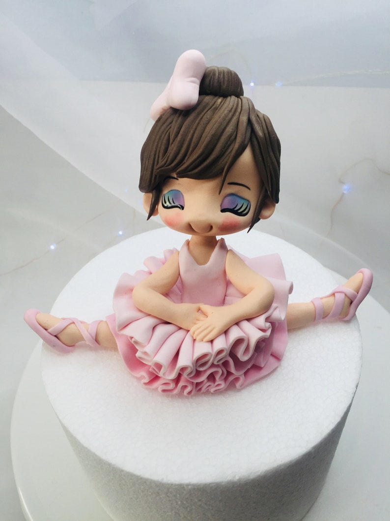 Puede incluir: Un adorno de pastel de bailarina de fondant rosa y marr&oacute;n con un lazo rosa en el pelo. Lleva un tut&uacute; rosa y zapatillas de ballet rosas.