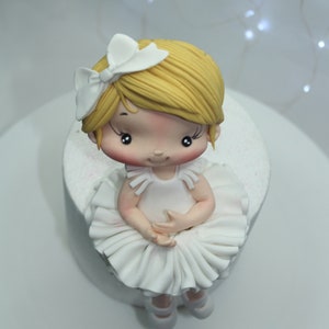 May include: A white fondant ballerina cake topper with a white bow, blonde hair, and a white tutu. The cake topper is sitting on a white cake.