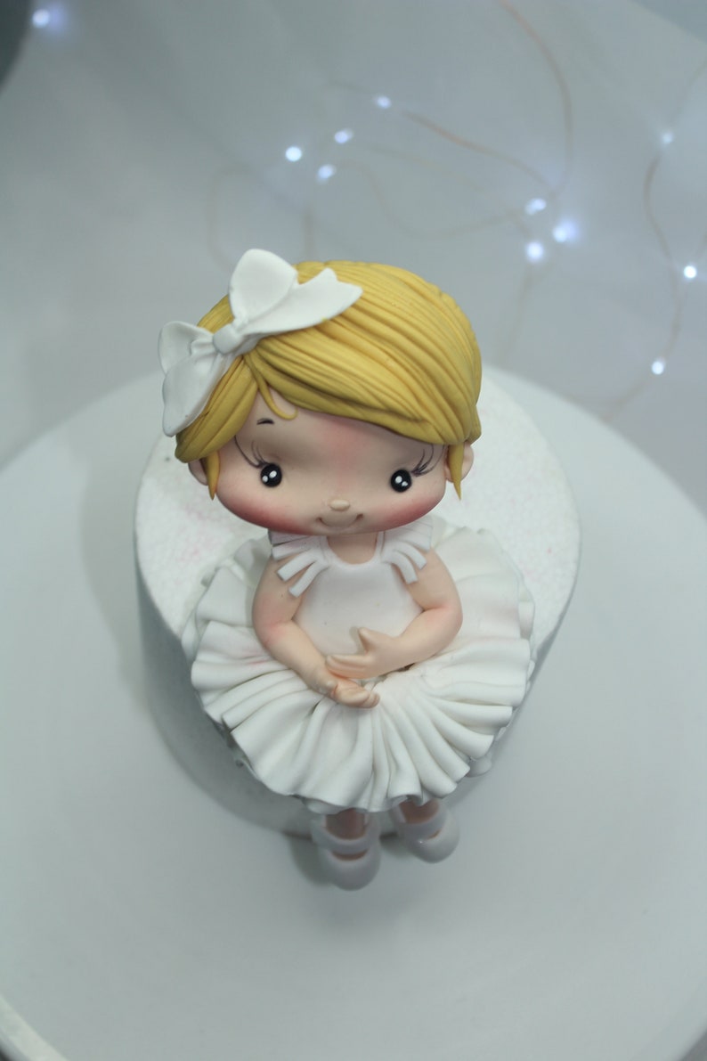 May include: A white fondant ballerina cake topper with a white bow, blonde hair, and a white tutu. The cake topper is sitting on a white cake.